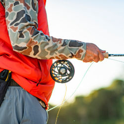 An angler wearing a camo-print long-sleeve and bright orange vest holds a Helios 3 rod with a Mirage reel.