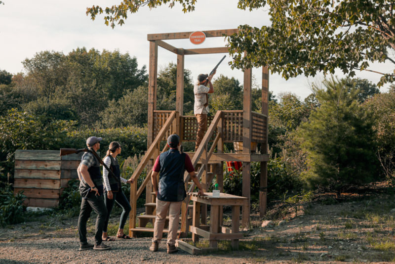 Several people gathered around a shooting stand watching a wingshooter shoot clays.