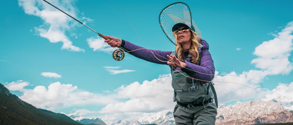 An angler in waders casts a fly rod surrounded by mountains and sky