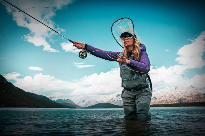 A woman casts line as she wades through a cold river with snowy mountains in the background.
