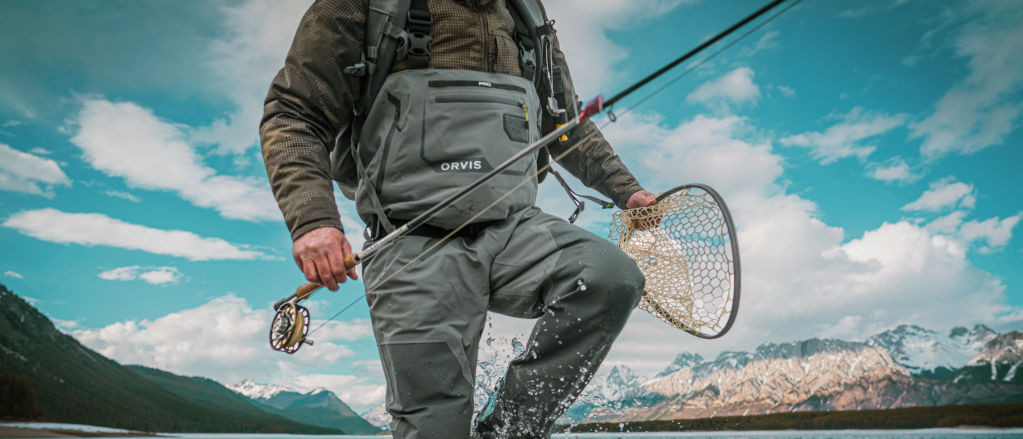 An angler wearing waders splashes through the shallow waters of a mountain lake.
