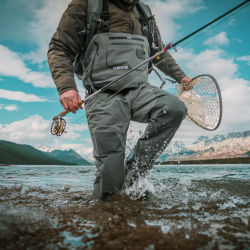 An angler wearing waders splashes through the shallow water of a mountain lake.