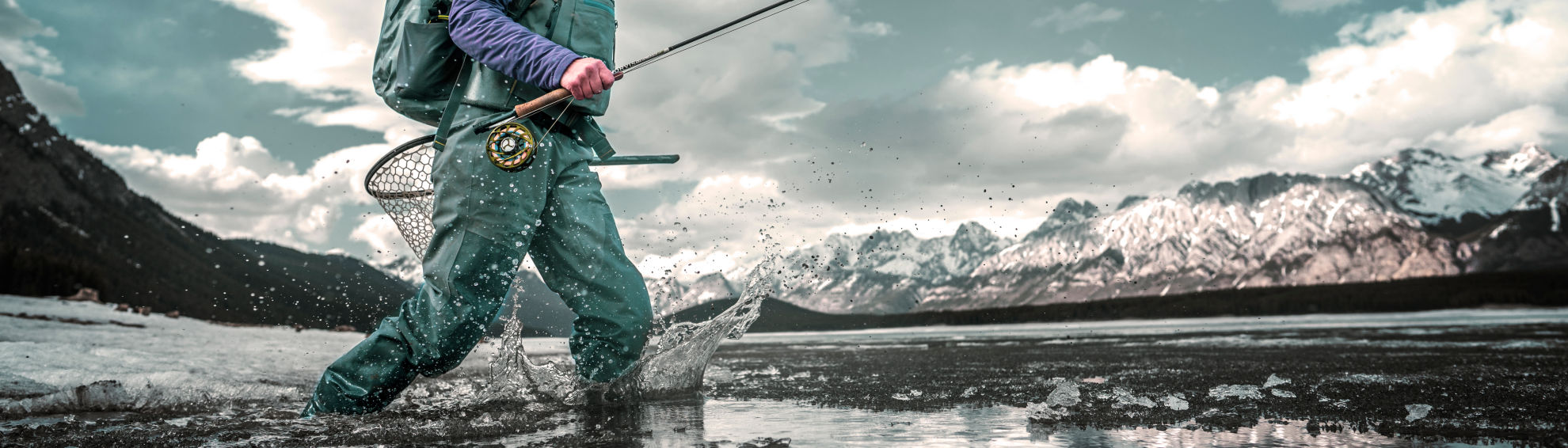 An angler in full waders and gear walking through a shallow river surrounded by snow-covered mountains