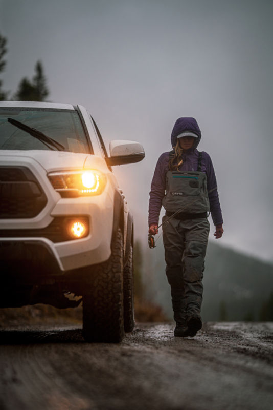 An angler in waders walking next to a truck.