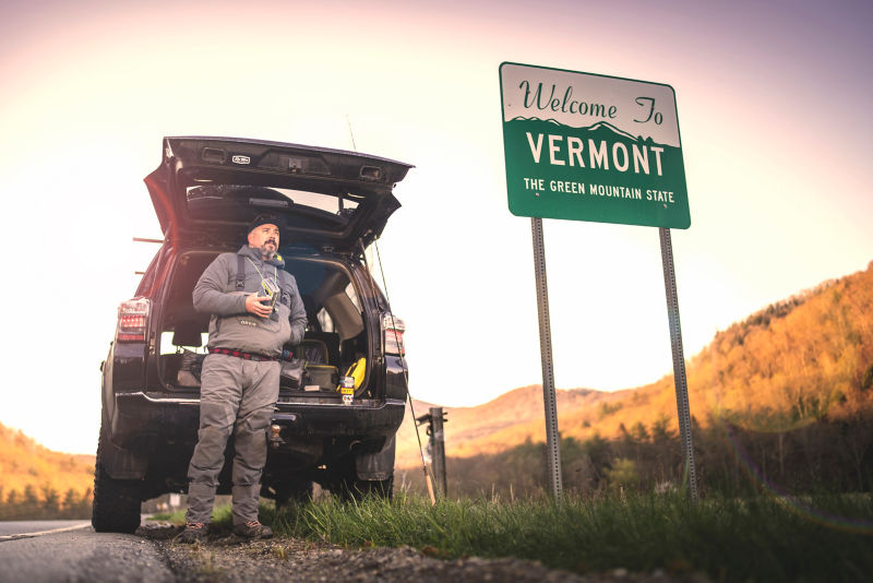 Man in full waders stands between the open hatch of an SUV and a Vermont state sign.