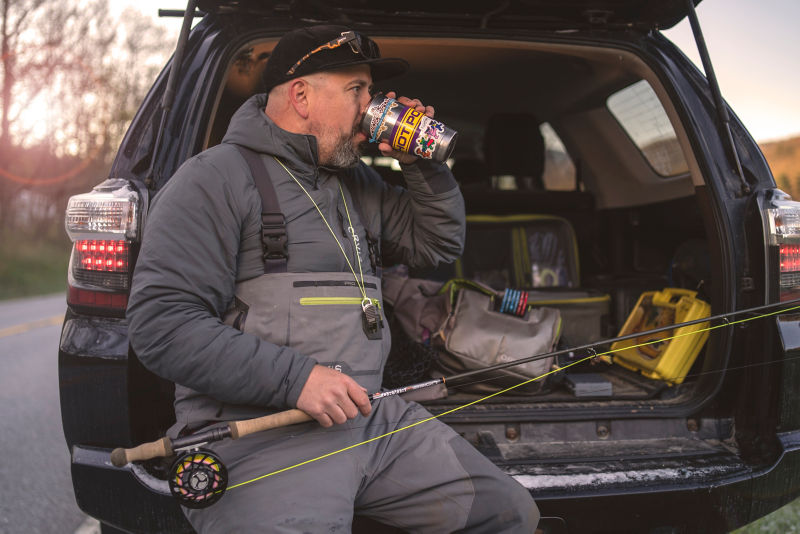 Man holding two-handed rod while sitting in the open hatch of an SUV.