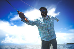 A sunglass-wearing angler with a buff pulled up over their face casts under sunny skies.