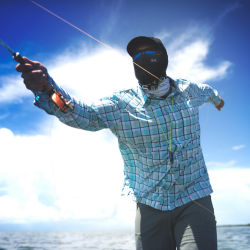 A sunglass-wearing angler with a buff pulled up over their face casts under sunny skies.