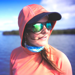 A woman wearing an orange shirt, hat, and sunglasses while near the water in the sun