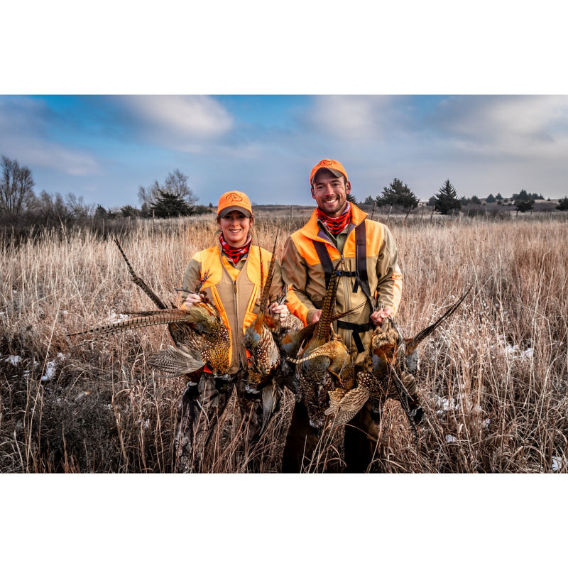 Ethan and Kat Pippitt stand in the midst of tall dry grass holding several pheasants.