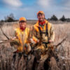 A woman and man wearing hunting gear standing in a field holding dead pheasants.