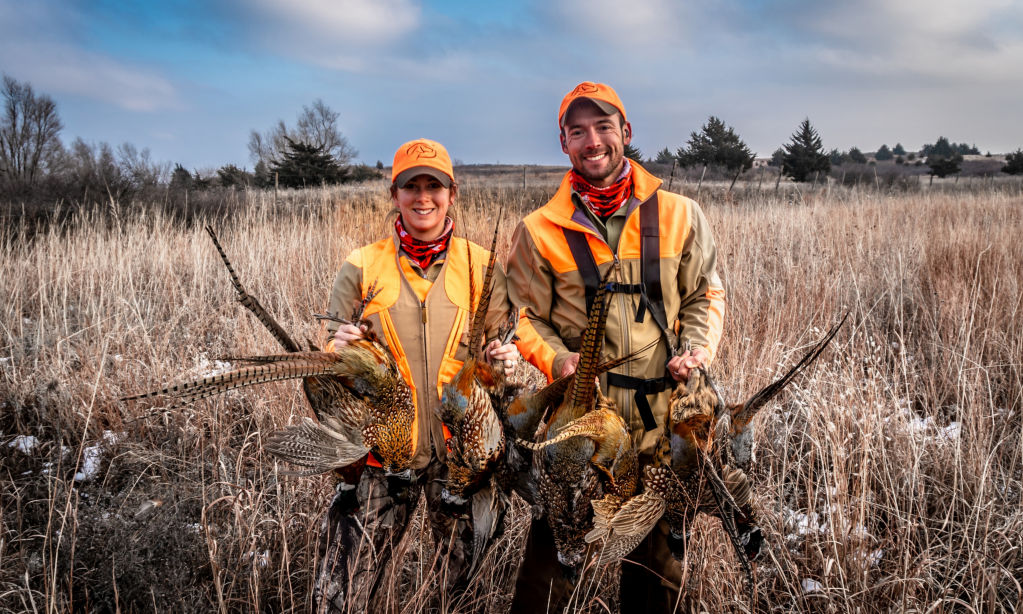 A woman and man wearing hunting gear standing in a field holding dead pheasants