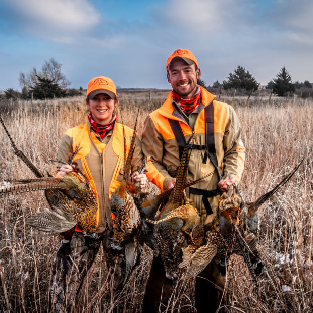 Ethan and Kat Pippitt stand in the midst of tall dry grass holding several pheasants.