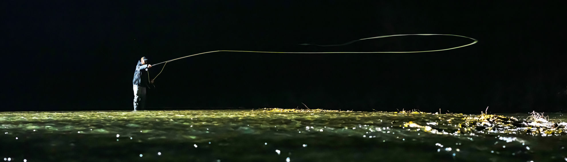 An angler casts across green waters in the dark of night.