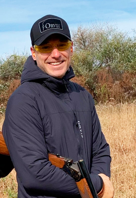 Robert Bean wearing shooting glasses smiles for the camera with a shotgun over his arm
