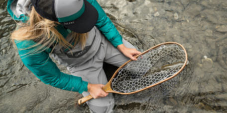 An angler wearing waders crouches in a shallow river, inspecting the fish in their net.