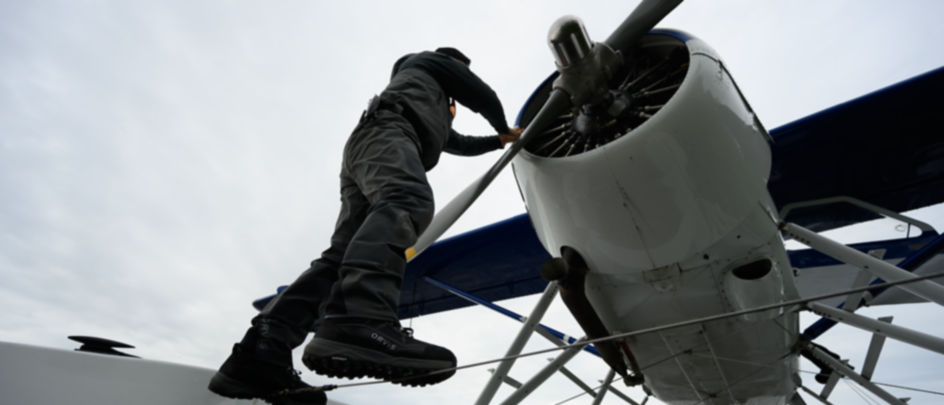 A man wearing waders balances on a cable while working on an airplane propeller.
