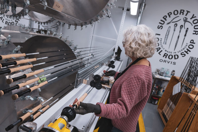 A woman works in the rod shop with a number of fly rods on a machine before her.