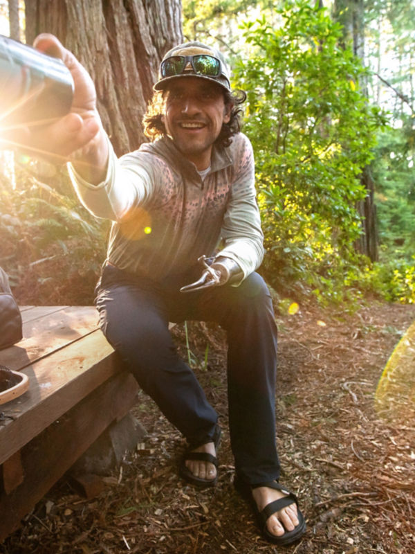 A hiker sits on a wooden deck offering a water bottle to someone off camera.