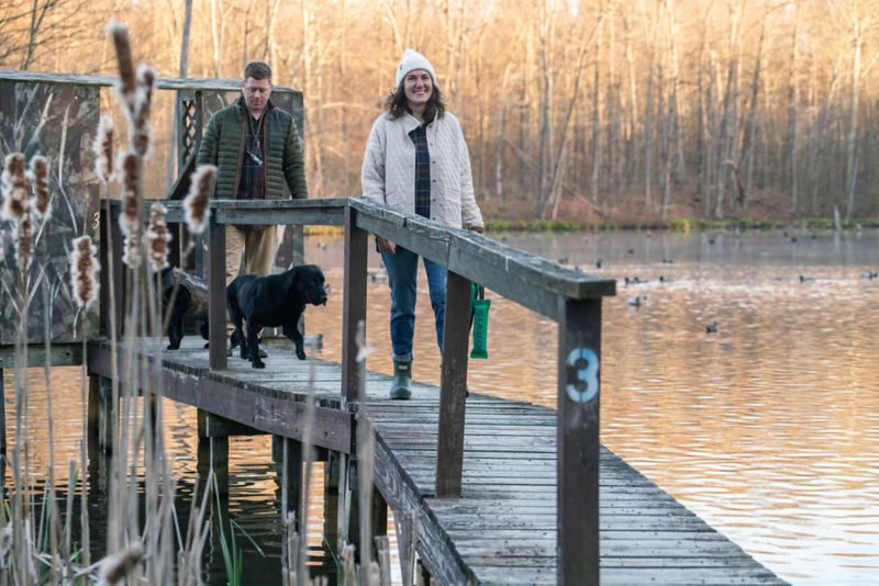 Two people and a black Labrador Retriever walk along a raised wooden walkway.