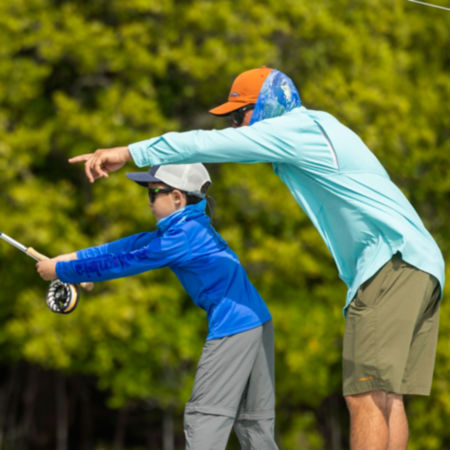 A man stands behind his son and guides him as he casts a fly rod.