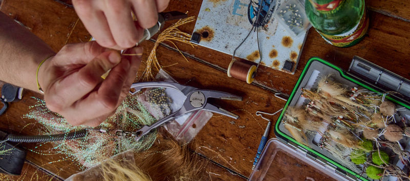 A close-up of two hands tying a fly with supplies on a wooden table.