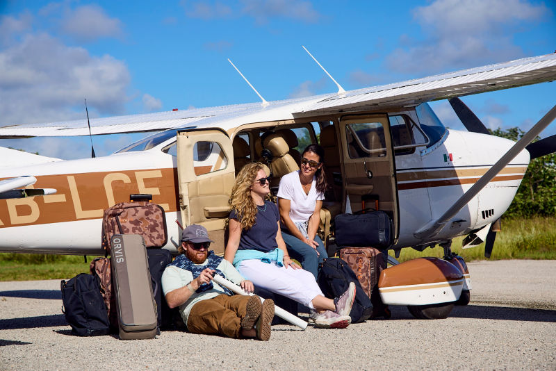 Three people wait next to a prop plane with their luggage.