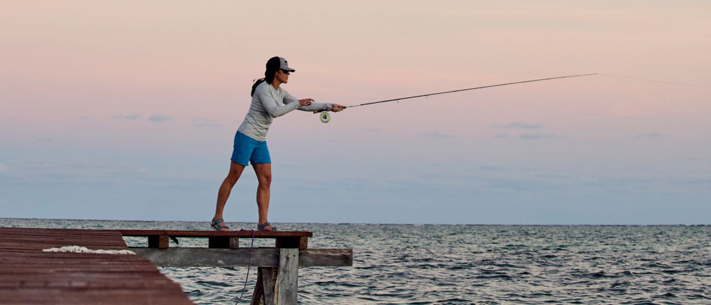 A woman standing at the end of a dock at sunset, casting a rod into the open water