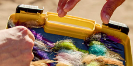 Two hands reach into a large fly box filled with brightly colored, fuzzy flies