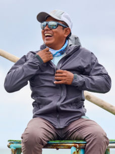 An angler smiles under a cloudy sky while adjusting his raincoat
