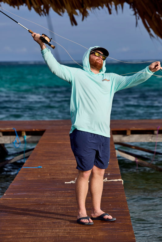 A man wearing a light green shirt, shorts and sunglasses casting into the ocean off of a dock