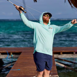 A man wearing a light green shirt, shorts and sunglasses casting into the ocean off of a dock