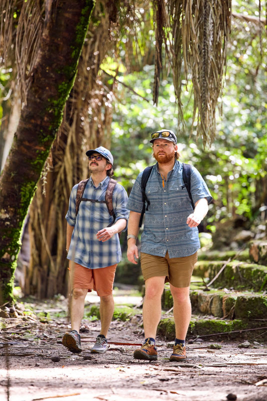 Two men wearing backpacks walking in a wooded, stoney area