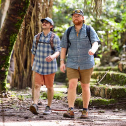 Two men wearing backpacks walking in a wooded, stoney area
