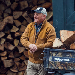 A man leans on a wagonload of wood next to a wood pile.