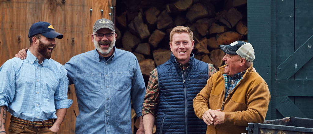 Four men laughing in front of a woodpile at Orvis Sandanona