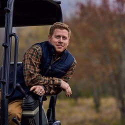 A man wearing a plaid shirt and blue vest leans out from the back of a golf cart.