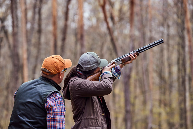 A hunter aims her shotgun with an instructor at her shoulder to offer guidance.