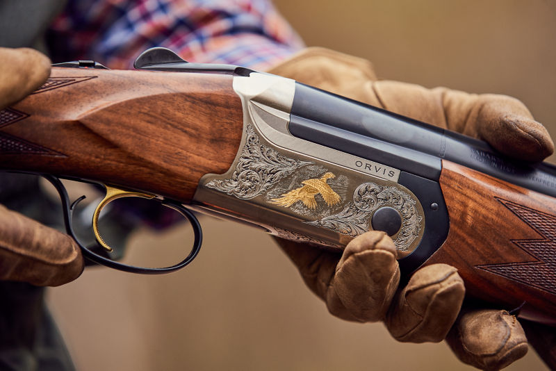 The intricate details of an Orvis ELOS over-under shotgun held in two leather-gloved hands.