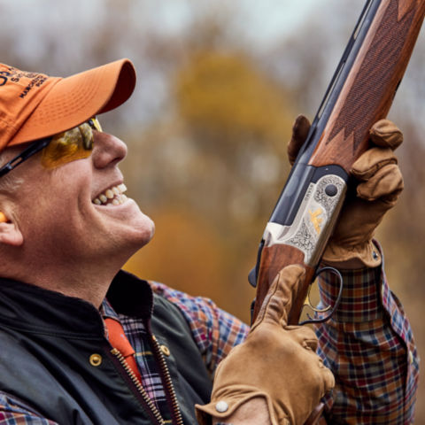 Man in orange hat holding shotgun smiling after shooting a sporting clay