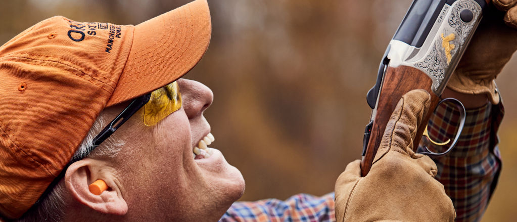 A hunter in an orange ball cap smiles as he aims his shotgun at the sky.
