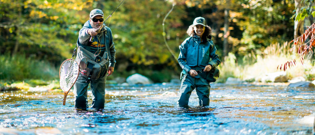A father casts his fly rod while his daughter watches from knee-deep in the stream.