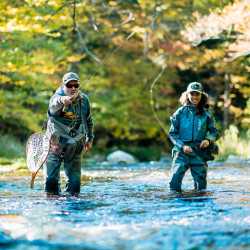 Two anglers in waders casting from knee-deep in a river.