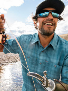 A man wearing a blue plaid short sleeved shirt, hat, and sunglasses casting a fly rod