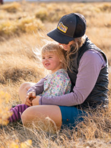 A person sits cross-legged in a field with a toddler in their lap
