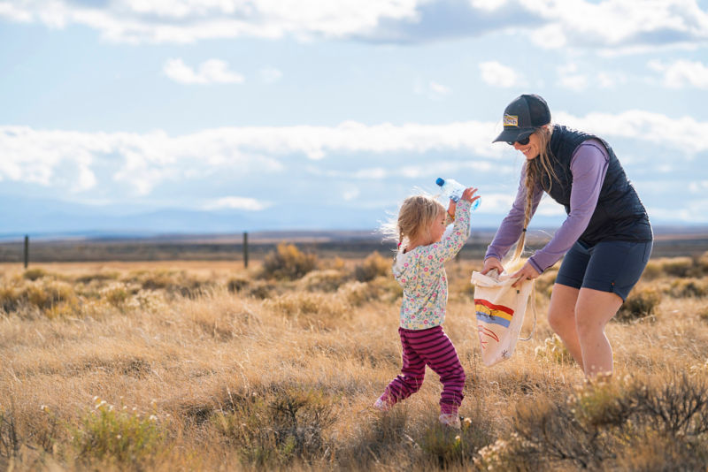 A woman in a field of dry grass holds a bag open for a child to put garbage in it.
