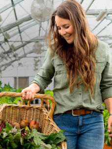 A woman carries a basket of vegetables in a greenhouse.