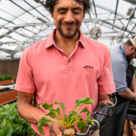 A man wearing a pink polo shirt in a greenhouse, holds a beet .