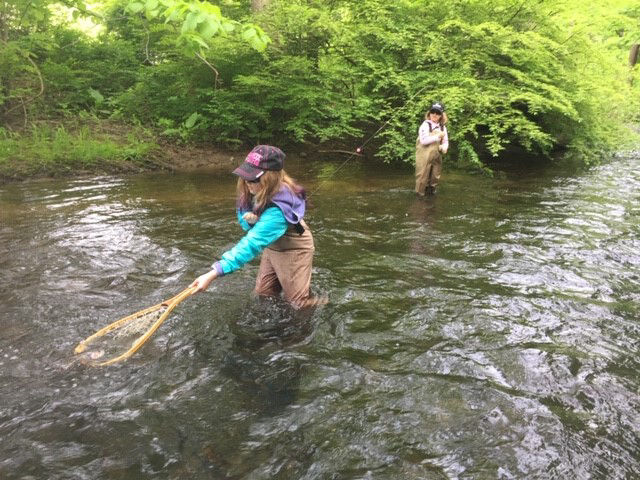 A girl wades in a stream to net a fish on the end of her sister's line.