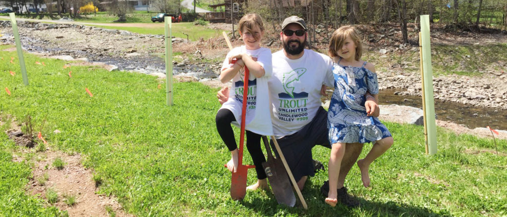 A smiling, bearded man poses with his two daughters near the stream.